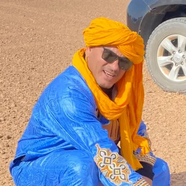 About us: Lahcen, founder and camp director, in a blue robe and orange turban in the desert