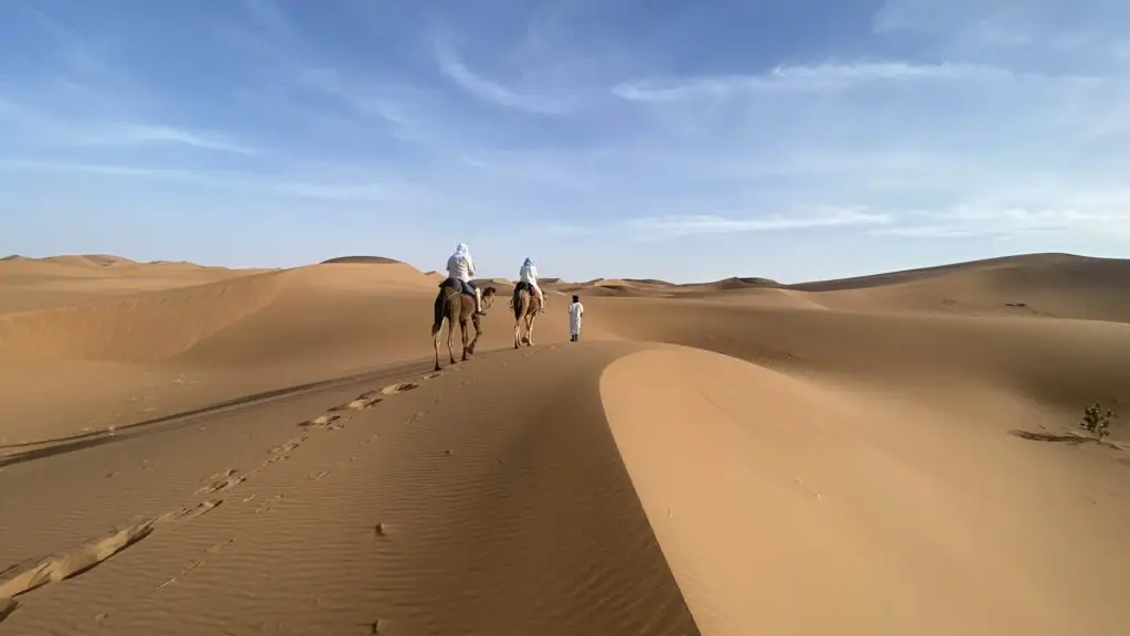 Two riders on camels and a guide on foot walking along a dune ridge during camel trekking